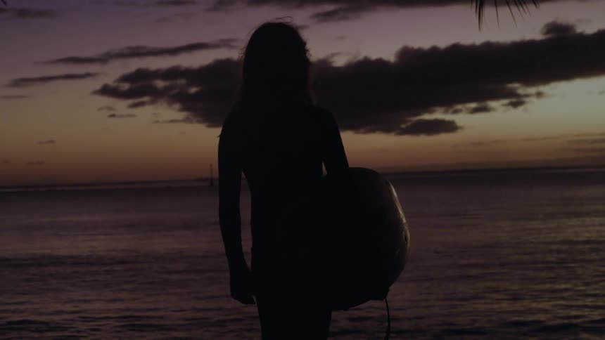 Silhouette of a female surfer holding her board looking out at the ocean during a beautiful sunset