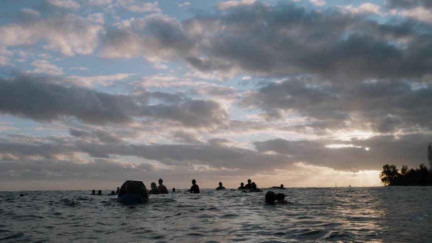 Silhouetted surfers wait in the calm ocean under a dramatic cloudy sky as the sun sets on the horizon