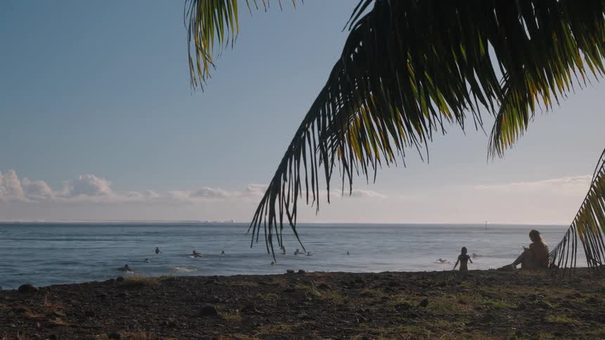 A mother and child relax on a tropical beach while surfers ride gentle waves under a palm tree at sunrise