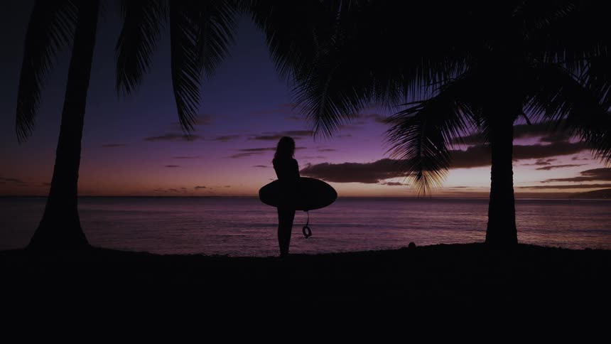 A female surfer stands with her surfboard on a tropical beach, silhouetted against a beautiful purple and orange sunset over the ocean