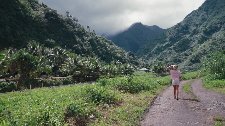A still shot of a young woman standing on a dirt road in a breathtaking tropical valley, looking up at the lush green mountains