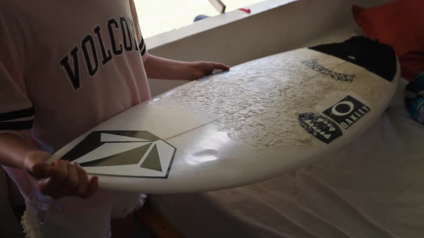 A close-up of a person in a pink shirt holding a white, waxed surfboard indoors