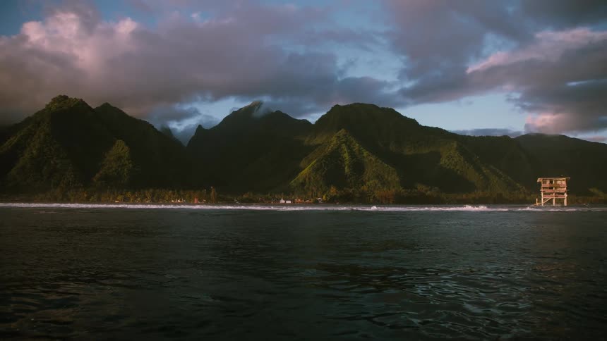 An epic wide shot of a tropical island coastline shows lush green mountains under dramatic clouds as waves break on the reef