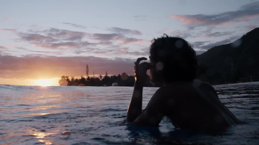 A young surfer sits on his board in the calm ocean, taking a quiet moment to enjoy the stunning tropical sunset