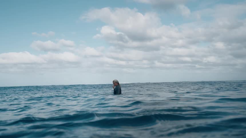 A serene slow-motion shot captures a woman in a wetsuit floating alone in the vast, open ocean while adjusting her snorkel