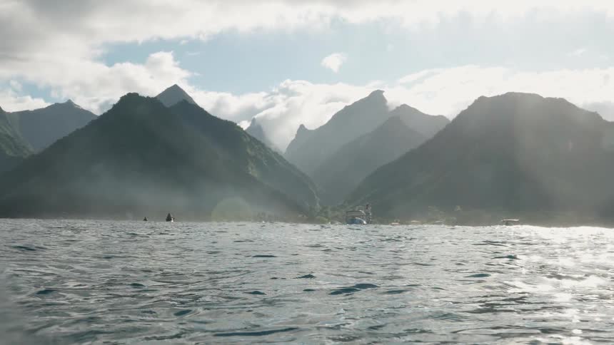 A view from the ocean's surface shows majestic, hazy green mountains on a tropical island under a bright, cloudy sky