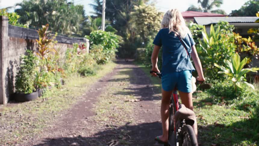 A young girl with blonde hair rides her bicycle down a sunny, tropical dirt path in slow motion