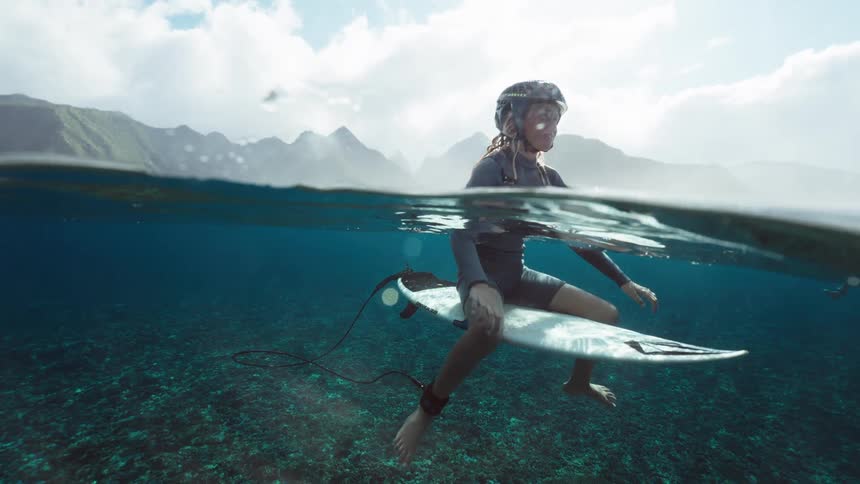 Stunning over-under shot of a young surfer waiting patiently on his board in the crystal clear tropical ocean with lush mountains in the distance