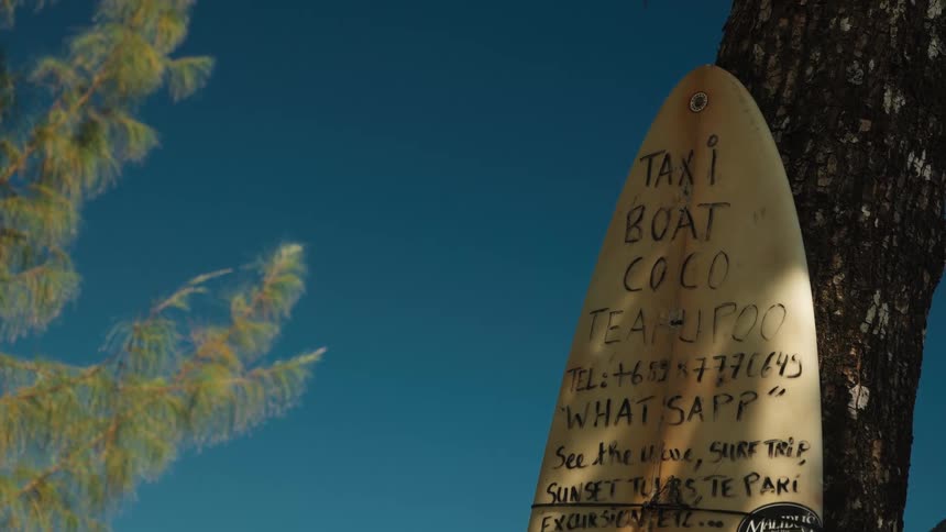 A surfboard with a handwritten advertisement for a taxi boat and surf trips in Teahupoo leans against a tree