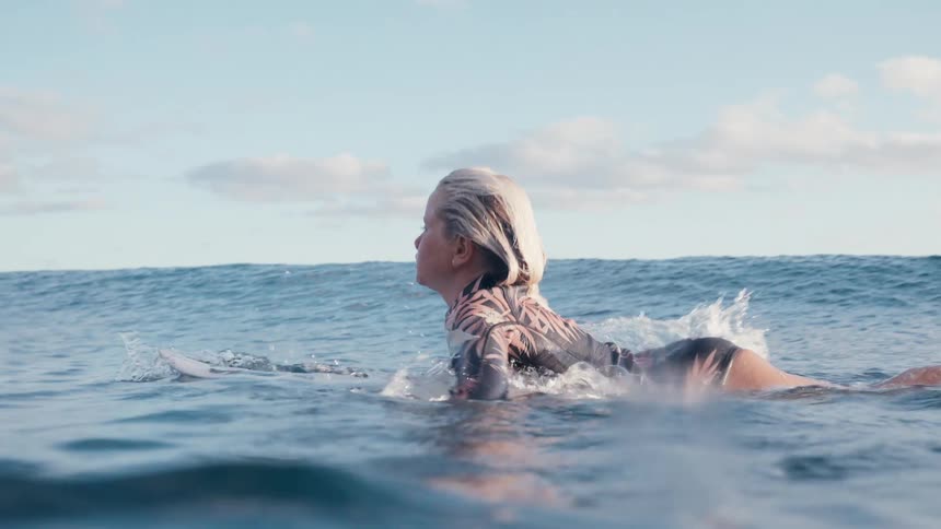 A young female surfer in a wetsuit paddles her surfboard through the calm ocean in beautiful slow motion