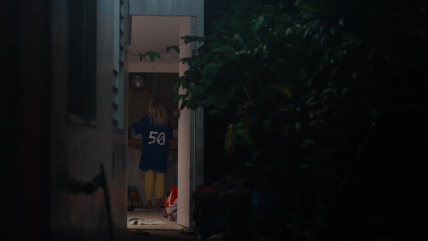 From a dark outdoor walkway, a blonde woman in an oversized jersey turns on the light and moves around in her simple kitchen at night