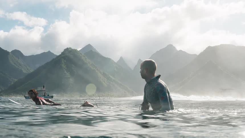 Surfers patiently wait in the ocean for the next wave, with a stunning backdrop of misty tropical mountains