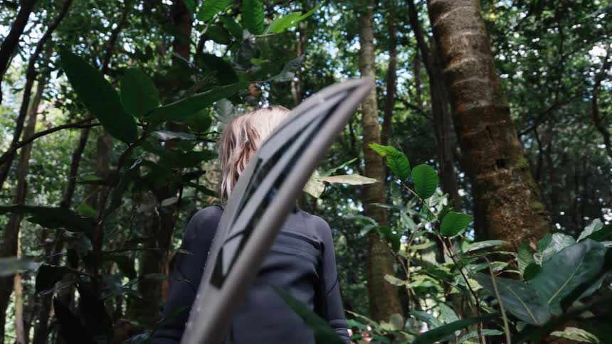 A young blonde surfer in a wetsuit stands with his board in a lush green jungle, looking up at the canopy