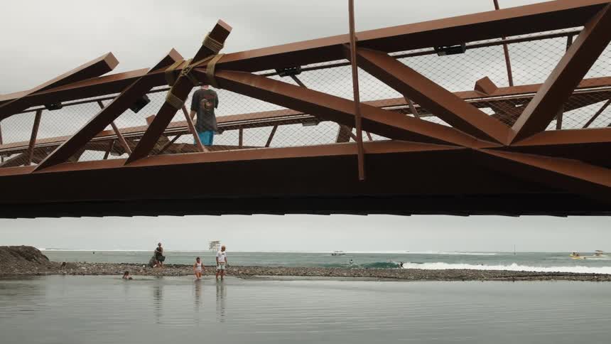 A modern architectural bridge frames a beach scene where people walk above, swim below, and surf on the ocean waves in the distance