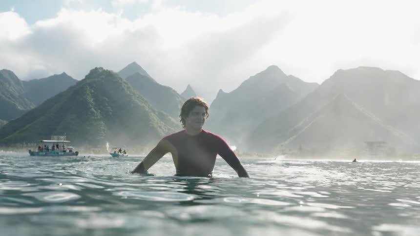 Slow motion shot of a male surfer sitting on his board in the ocean, smiling with beautiful lush mountains in the background