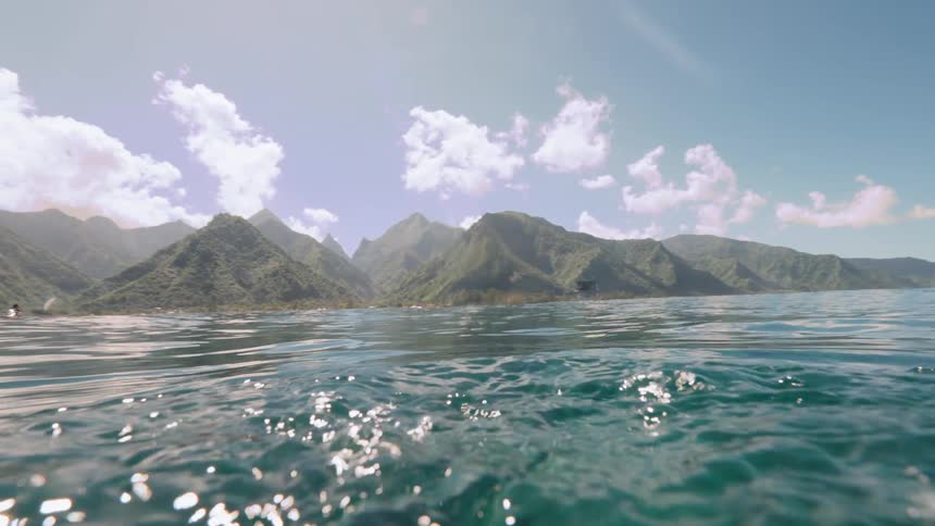 A slow-motion shot emerges from underwater to reveal a stunning tropical island with lush green mountains under a bright blue sky