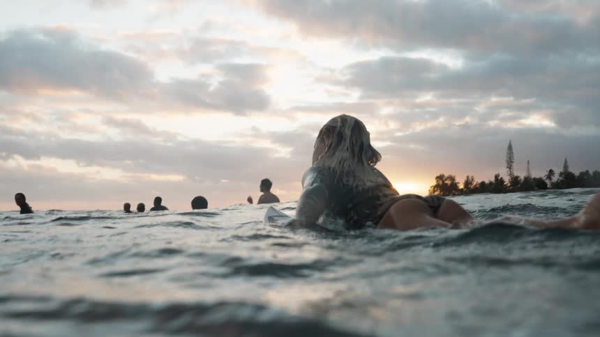 Slow motion over-the-shoulder shot of a female surfer paddling on her board in the ocean during a beautiful sunset