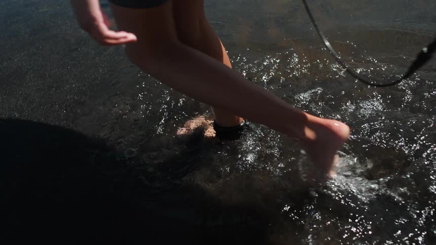 A surfer with a leash on their ankle walks into the dark, shallow water of the ocean in slow motion