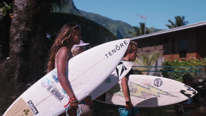 A group of cool young surfers carry their boards while walking through a sunny tropical town in slow motion