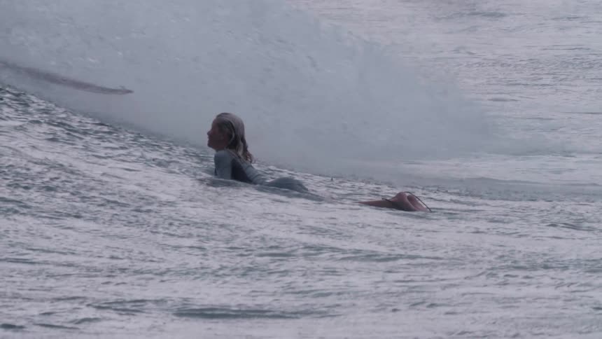 Slow motion side view of a female surfer paddling on her board as a large ocean wave rolls underneath her