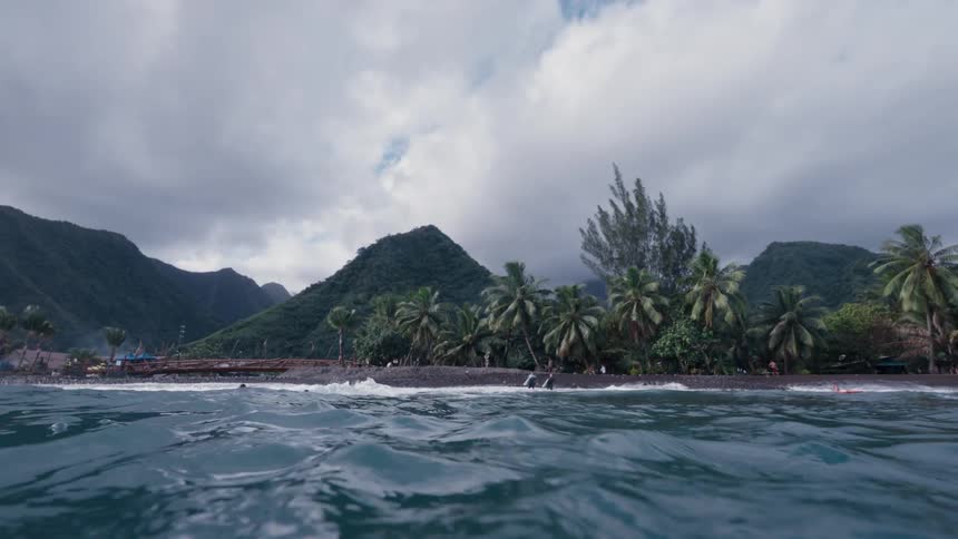 An immersive split-level view emerges from the turquoise ocean to reveal a stunning tropical beach with lush mountains under a cloudy sky