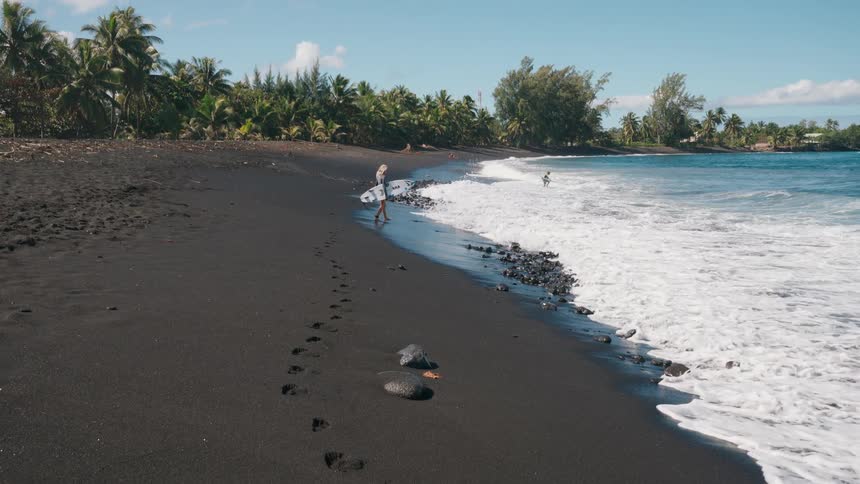 Footprints in the black volcanic sand lead to a surfer girl walking along the tropical shoreline with her board