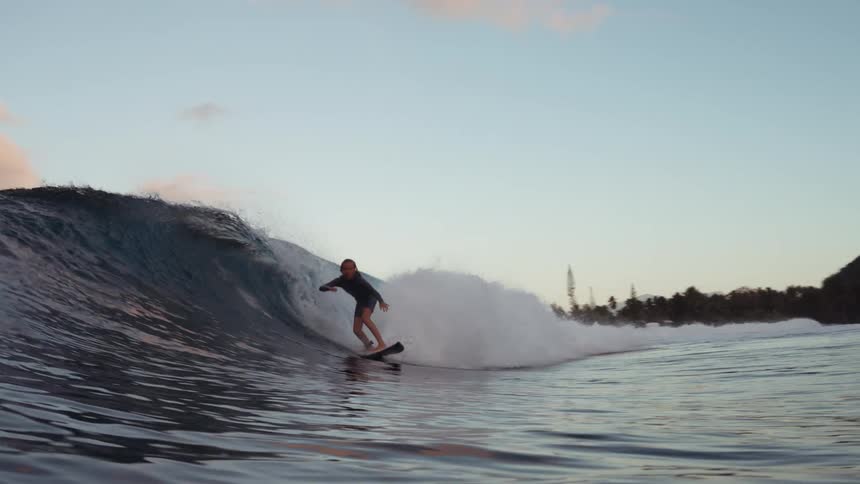 A young surfer skillfully rides a beautiful blue wave in slow motion at sunset on a tropical coastline