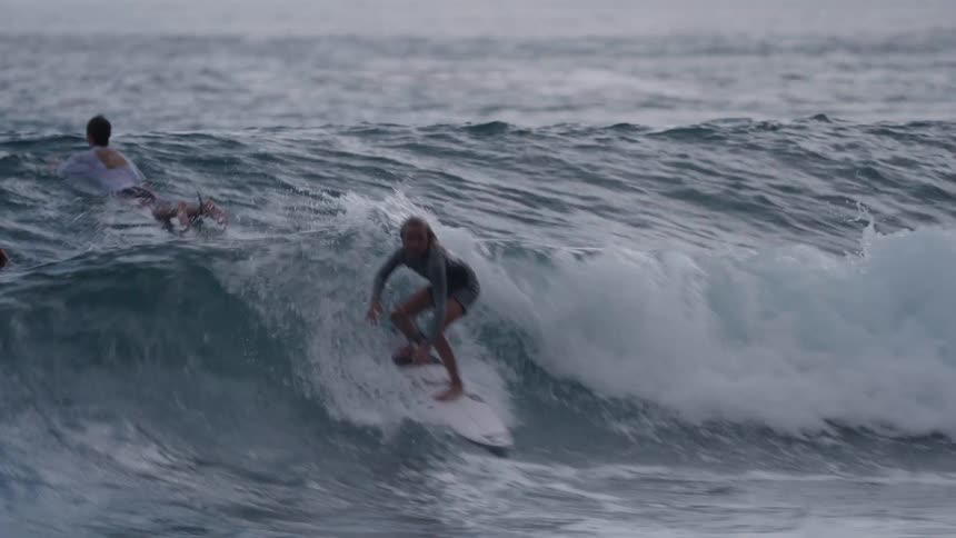 A young female surfer paddles to catch a wave, pops up on her board, and expertly rides it in slow motion