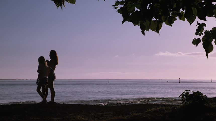 Two silhouetted young girls stand on a beach looking out at the calm ocean under a beautiful pastel sky