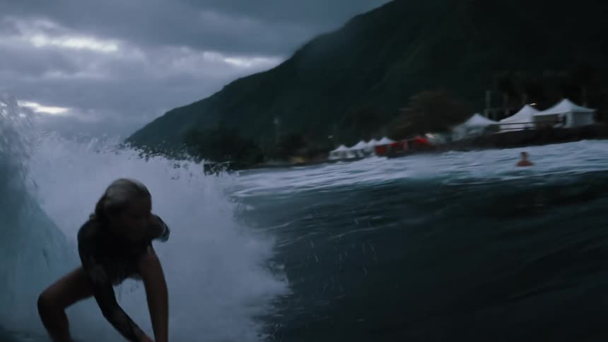 Immersive slow-motion water shot follows a female surfer riding a massive ocean wave towards a mountainous shore