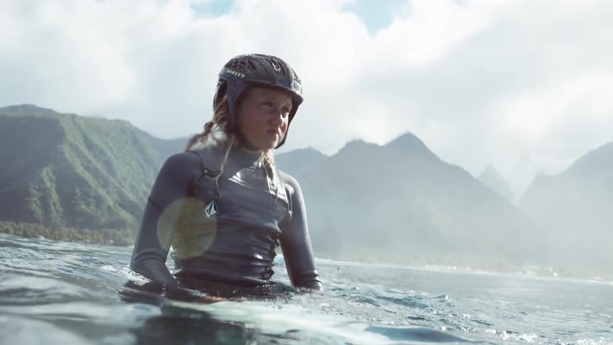 A determined young female surfer wearing a helmet and wetsuit sits on her board in the beautiful ocean, with dramatic mountains in the background
