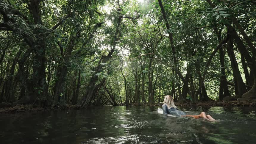 A female surfer paddles her board down a calm river through a dense tropical jungle
