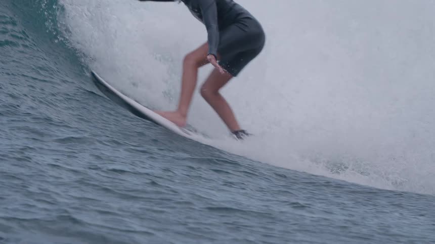 A determined female surfer in a wetsuit skillfully rides a powerful ocean wave in slow motion, carving through the water with precision