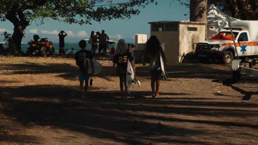 Three young friends with surfboards walk barefoot along a shaded, dusty path towards a sunny beach to go surfing