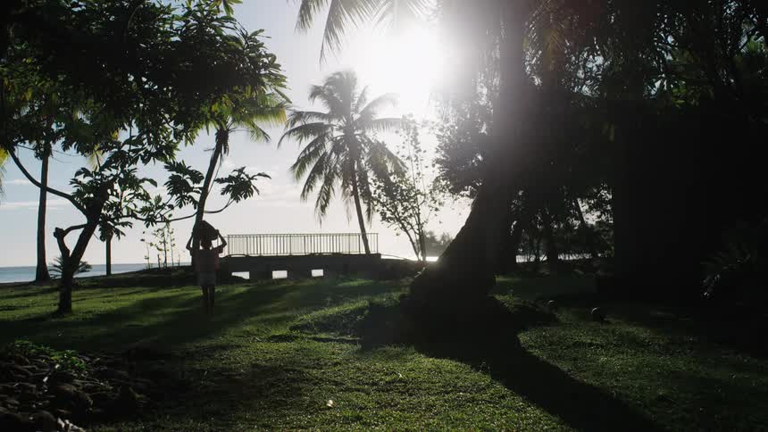 A young female surfer carries her surfboard through a tropical, sun-drenched park with palm trees