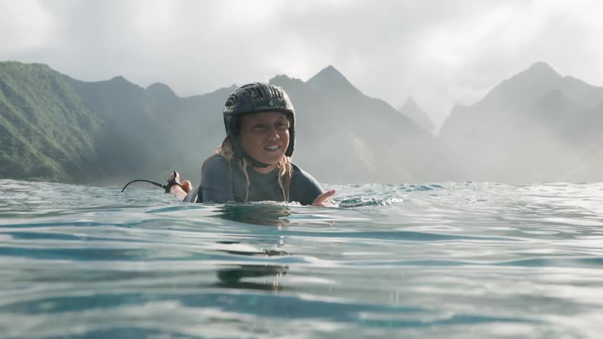 Slow motion shot of a happy female surfer paddling on her board in the calm ocean with a beautiful tropical mountain landscape in the background