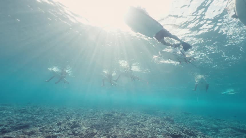 An underwater low-angle shot of a group of surfers waiting for waves as beautiful sun rays pierce the clear blue ocean water