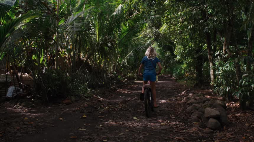 A young woman with blonde hair rides her bicycle down a lush, green jungle path in slow motion