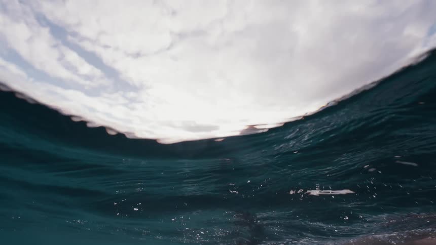 Slow motion split-level view transitions from underwater to show a female surfer paddling on her board in the tranquil ocean