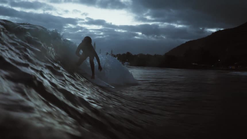 A silhouetted surfer expertly carves a dark ocean wave in stunning slow motion under a moody, overcast sky