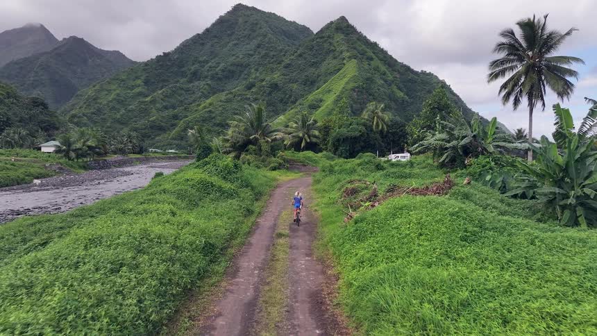 A person cycles down a dirt path through a lush green valley with majestic tropical mountains in the background