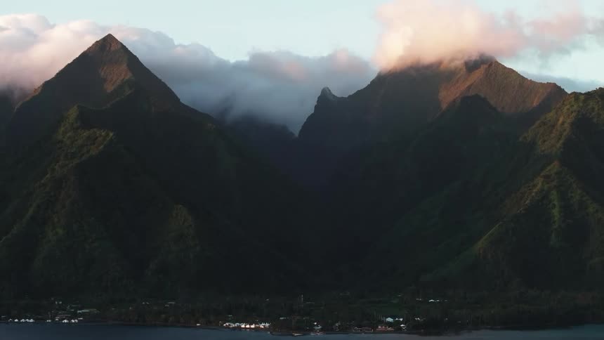 A static wide shot captures the majestic, cloud-capped mountains of Tahiti illuminated by the golden morning sun, overlooking a serene coastal town