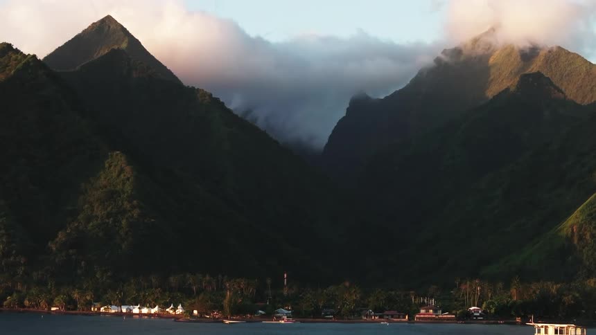 A serene view of a tropical coastal town nestled at the base of majestic green mountains, with soft clouds filling the valley at sunrise
