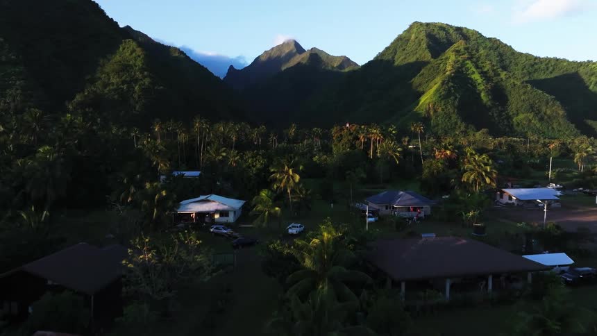 A slow aerial push-in over a tranquil tropical village in a lush valley, with majestic green mountains illuminated by the morning sun