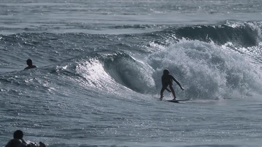 A silhouetted surfer skillfully catches and rides a powerful ocean wave in dramatic slow motion, carving a turn and sending spray flying