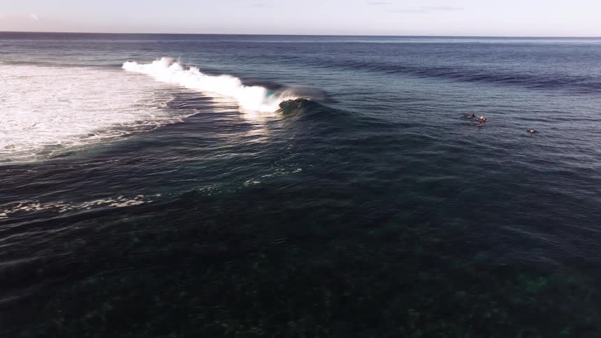 A majestic ocean wave breaks in slow motion as seen from an aerial perspective, with a group of surfers waiting nearby