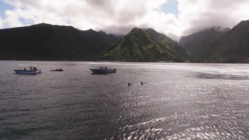 Aerial footage pans across the vast ocean, revealing numerous spectator boats and surfers gathered near a tropical island with lush green mountains