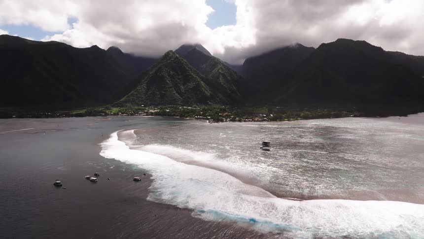 A stunning aerial view reveals the powerful waves of the Teahupo'o surf break against the dramatic mountainous coastline of Tahiti