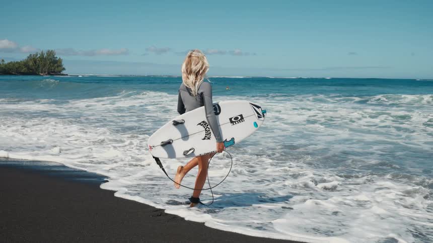 A female surfer in a wetsuit holds her board on a black sand beach, watching the waves roll in