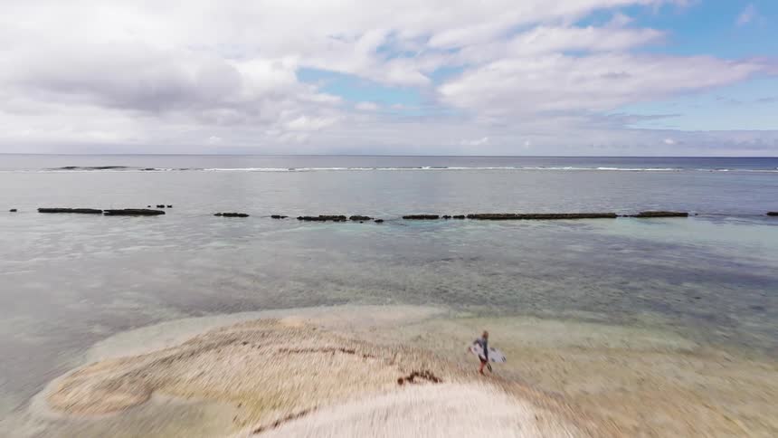 An epic aerial reveals a lone female surfer with her board on a remote sandy islet surrounded by a calm tropical ocean